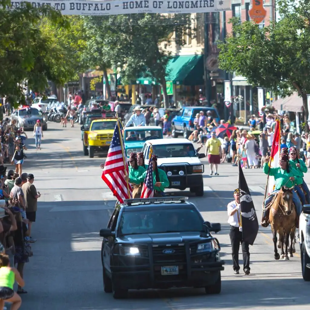 A police car, riders on horseback bolding flags, and a series of vehicles cruise north on Main Street during the annual Longmire Days parade under a banner stretched over the street says, “Welcome to Buffalo – Home of Longmire.”
