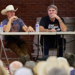 Derek Phillips and Robert Taylor, actors in the Longmire television series, hold microphones while making a presentation seated at a table on a stage with a full audience in the foreground at the Bomber Mountain Civic Center gymnasium in 2023.