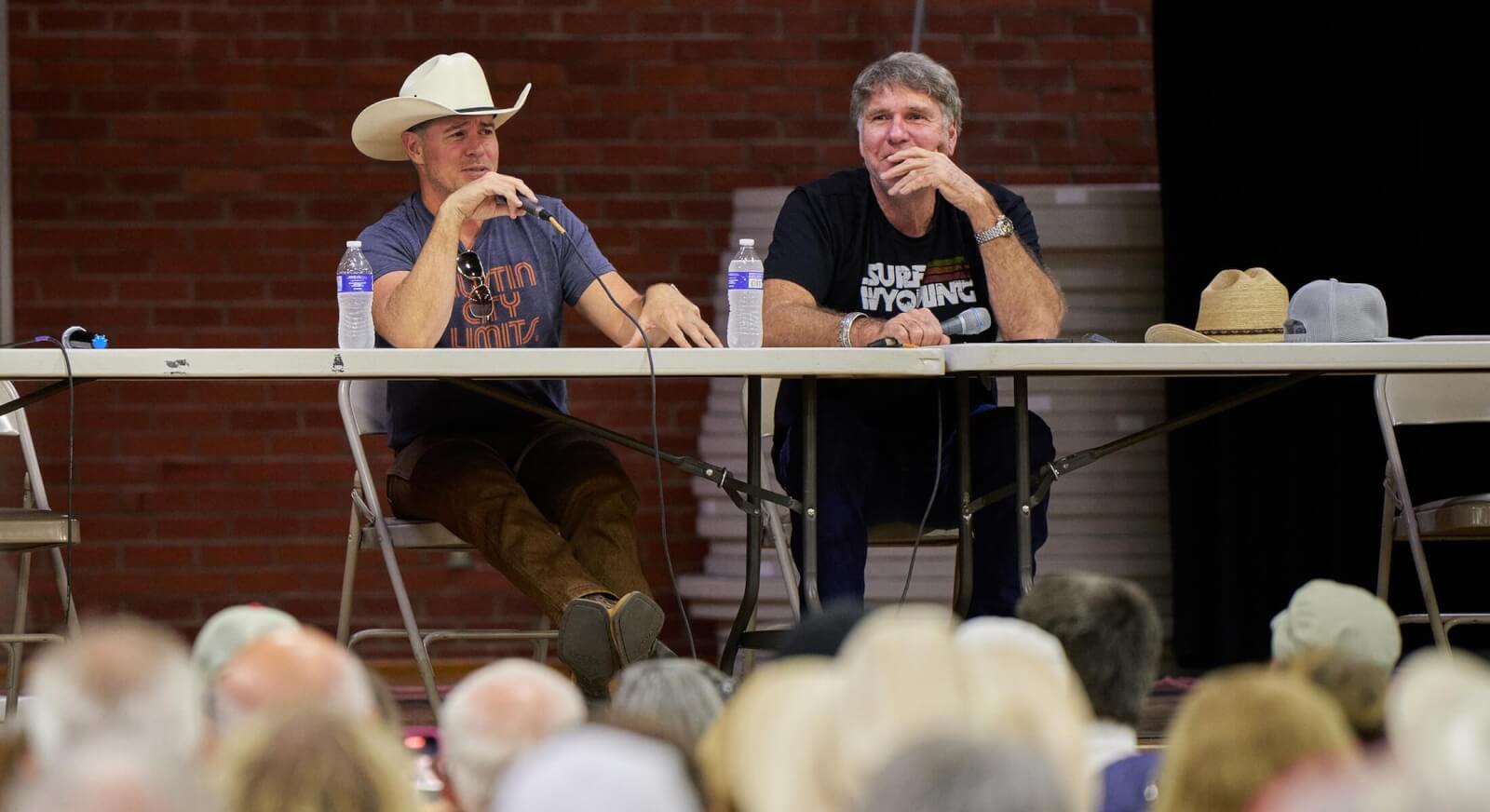 Derek Phillips and Robert Taylor, actors in the Longmire television series, hold microphones while making a presentation seated at a table on a stage with a full audience in the foreground at the Bomber Mountain Civic Center gymnasium in 2023.
