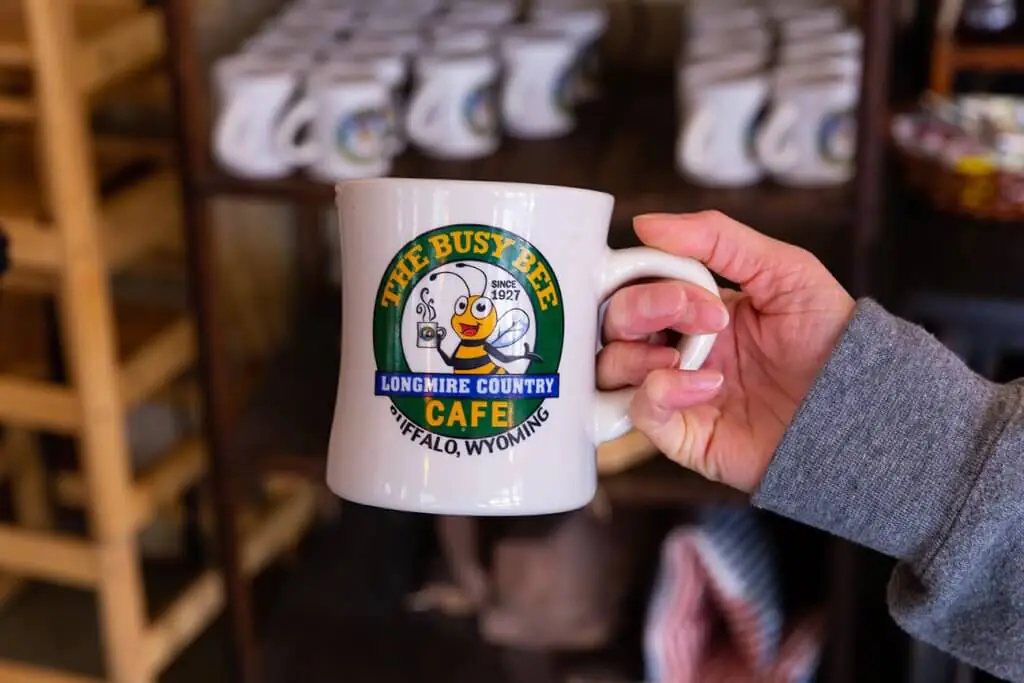 A patron holds a white coffee mug from the Busy Bee Cafe with a bee drinking coffee logo that says Since 1927, Longmire Country, Buffalo, Wyoming.