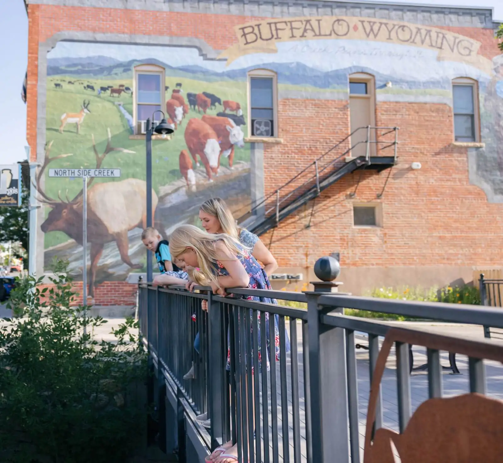 A mother and children looking over a railing in downtown Buffalo, WY, with a colorful mural in the background, showcasing one of the top places to visit in Buffalo, WY.