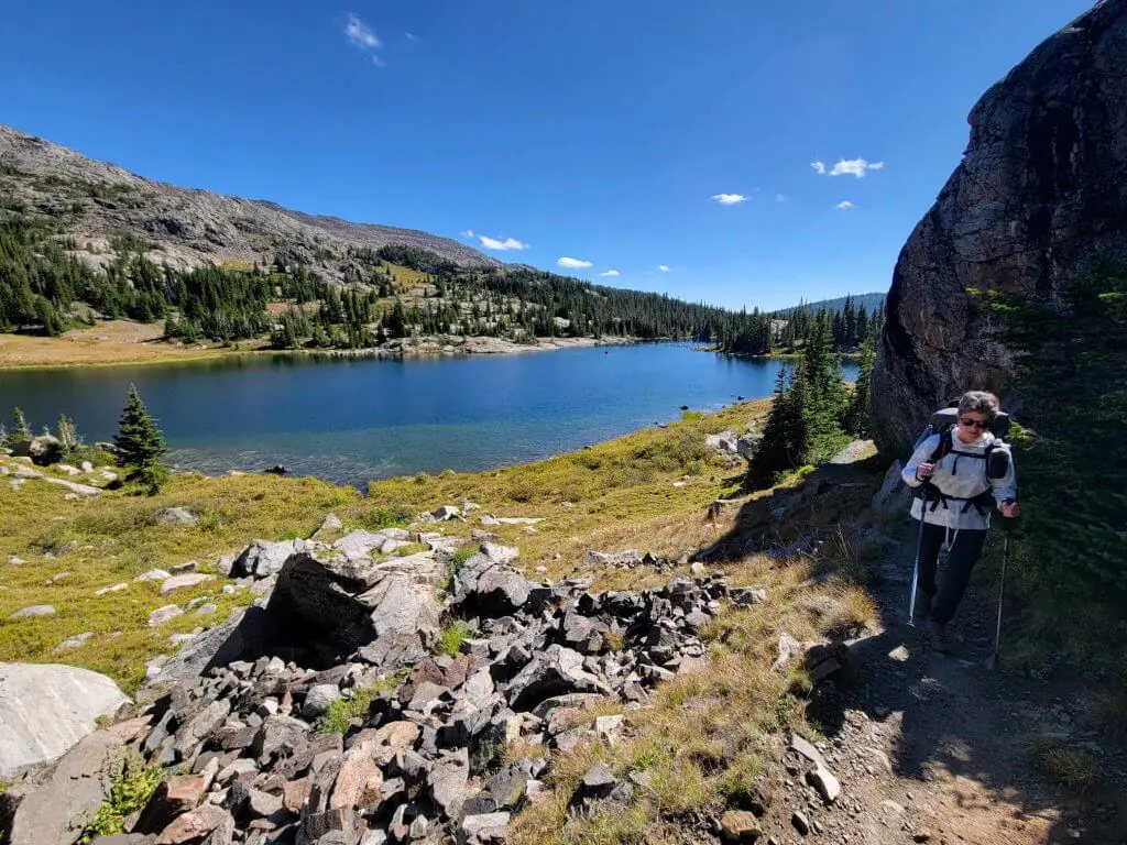 A hiker with a backpack trekking along a rocky trail beside Bighorn Lake, surrounded by lush mountain scenery on a sunny day.