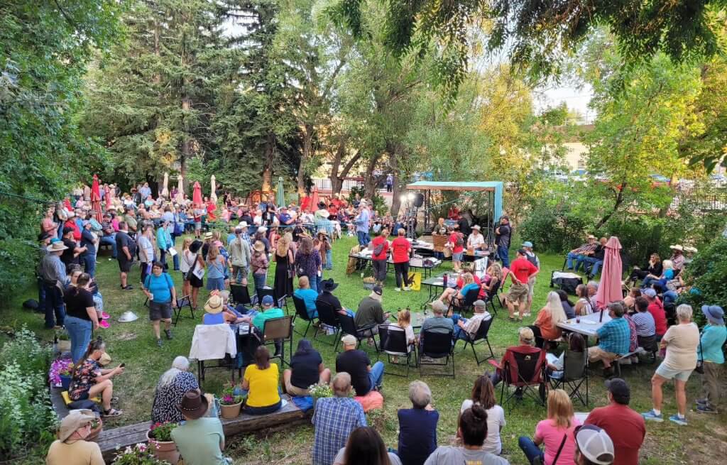 Outdoor event at the Occidental Hotel backyard in Buffalo, Wyoming, with a vibrant crowd gathered in a lush garden setting for a community event.