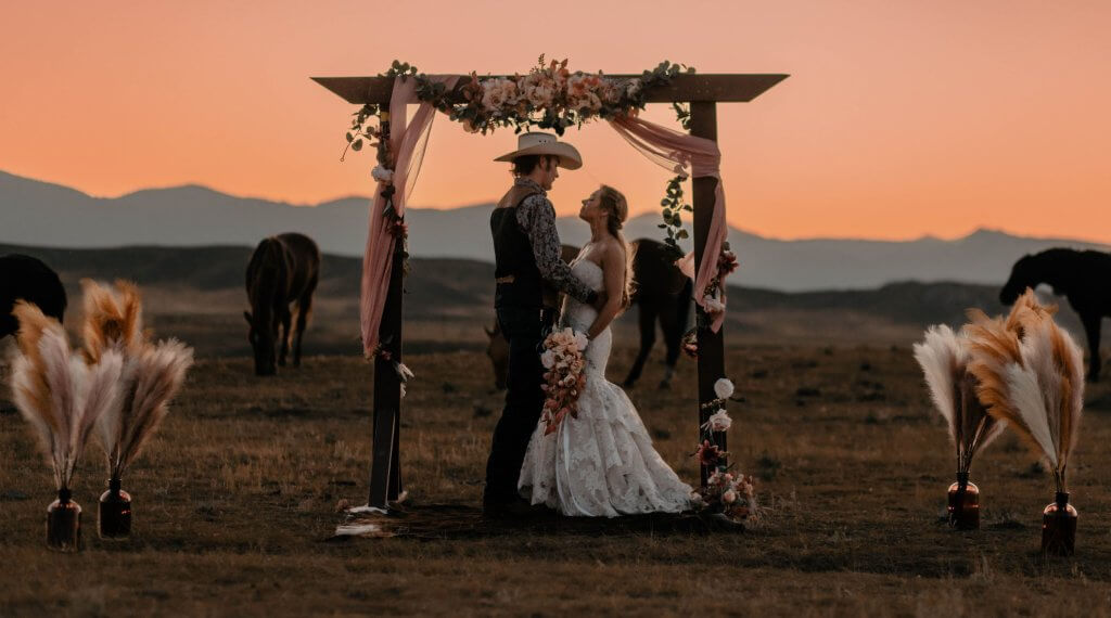 Scenic outdoor wedding venue at TA Ranch in Buffalo, Wyoming, with a couple under a floral arch at sunset surrounded by a rustic prairie and horses.