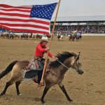 A cowgirl carries the American flag on horseback during the grand entry at the Johnson County Fairgrounds rodeo in Buffalo, WY, with a crowd and other riders in the background.