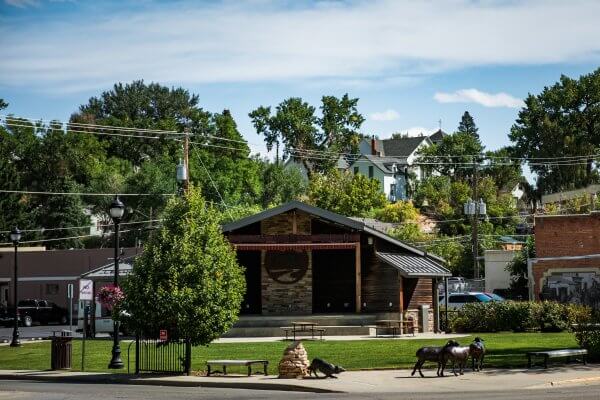 20 Free and Budget-Friendly Things to Do in Buffalo, Wyoming Crazy Woman Square in Buffalo, Wyoming, featuring a wooden stage with picnic tables, green lawn, benches, and bronze livestock sculptures under a partly cloudy sky.