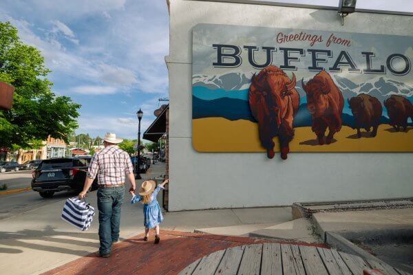 20 Free and Budget-Friendly Things to Do in Buffalo, Wyoming Man and young girl in cowboy hats walking hand-in-hand past a large mural that reads “Greetings from Buffalo”