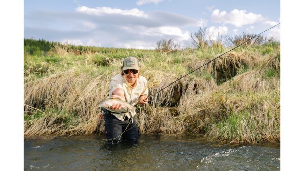 20 Free and Budget-Friendly Things to Do in Buffalo, Wyoming Smiling angler fly fishing in a creek near Buffalo, Wyoming, holding a freshly caught trout in front of grassy banks under a partly cloudy sky.