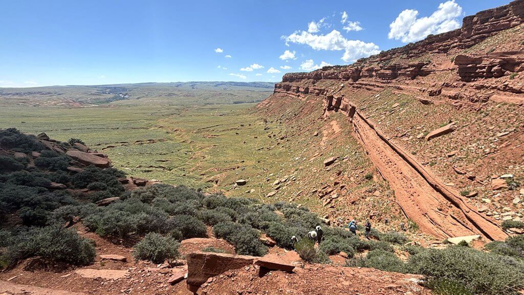 Hole in the Wall Hikers descending the Hole-in-the-Wall Trail in Wyoming’s Red Desert, with sweeping views of red rock cliffs and expansive green valley under a bright blue sky