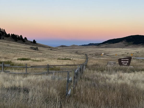 20 Free and Budget-Friendly Things to Do in Buffalo, Wyoming Sunset over rolling hills and open grasslands near the entrance to Bighorn National Forest west of Buffalo, Wyoming, with a wooden fence and forest sign in the foreground.
