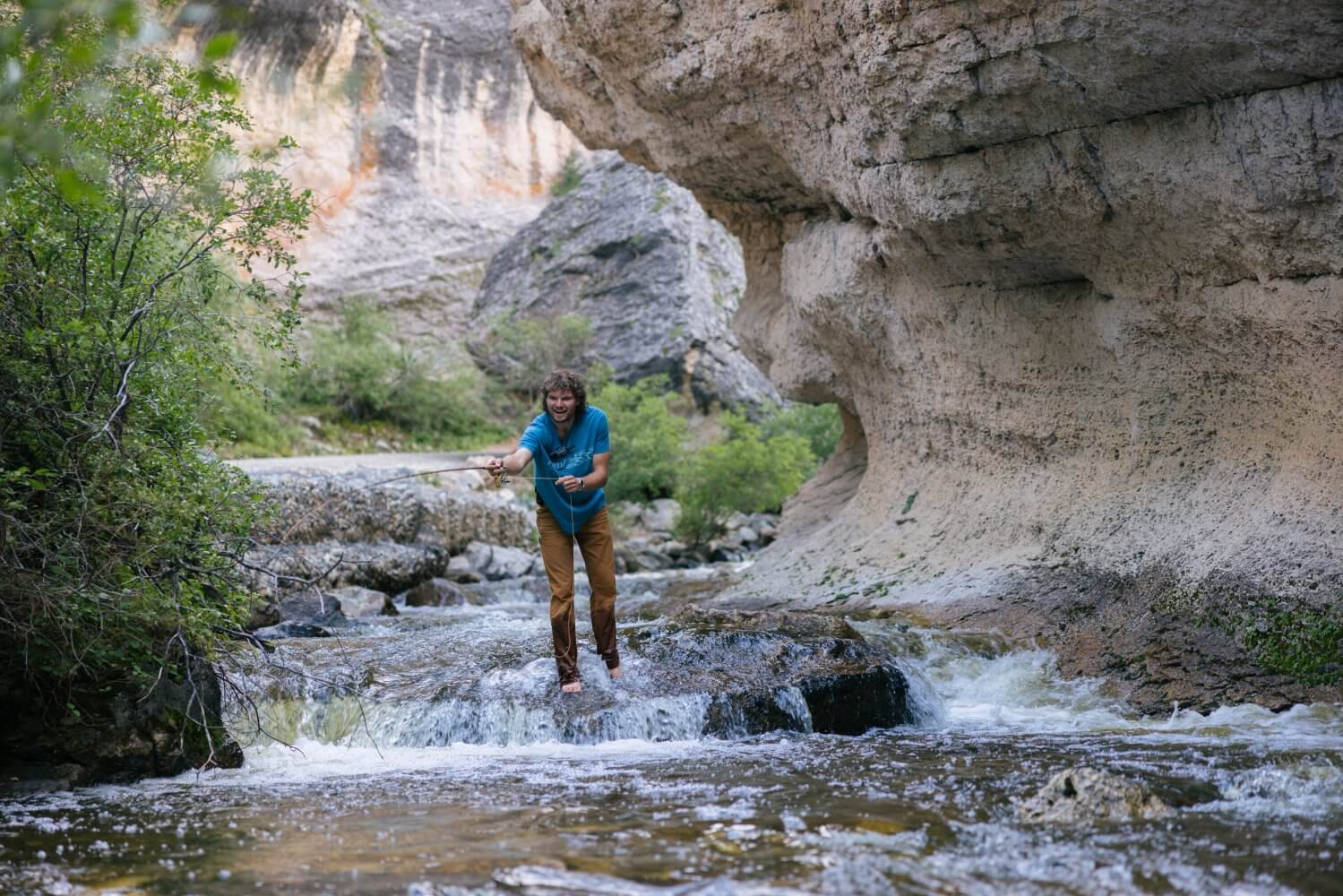 Origins of Crazy Woman Canyon Man fly fishing barefoot in Crazy Woman Creek beneath carved canyon walls, surrounded by wild rock formations and forest.