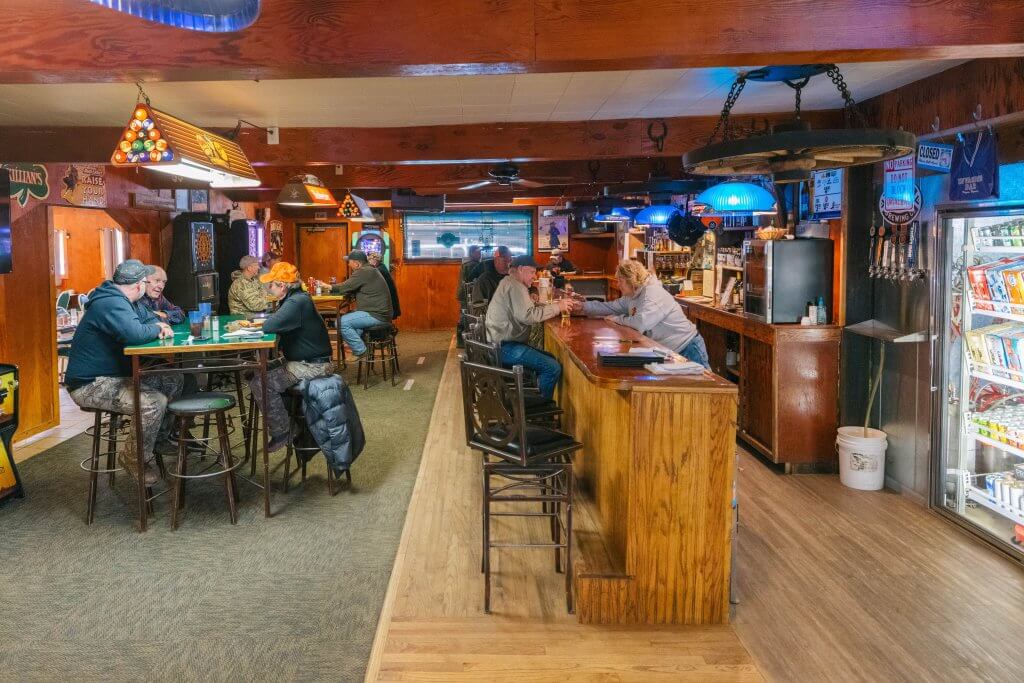 Hole in the Wall Invasion Bar & Restaurant interior showing wooden bar with patrons, vintage beer signs, pool table lighting, and authentic western saloon décor